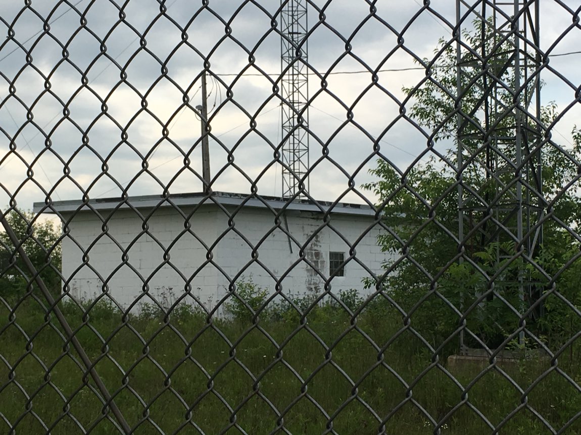 A white cinder-block building and a guyed steel tower seen through a chain-link fence topped with barbed wire